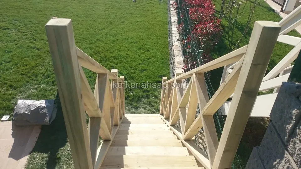 Yeşil çimliğe inen ahşap basamakların iki yanında kalın direkli, çapraz çıtalı açık tonlu korkuluk
