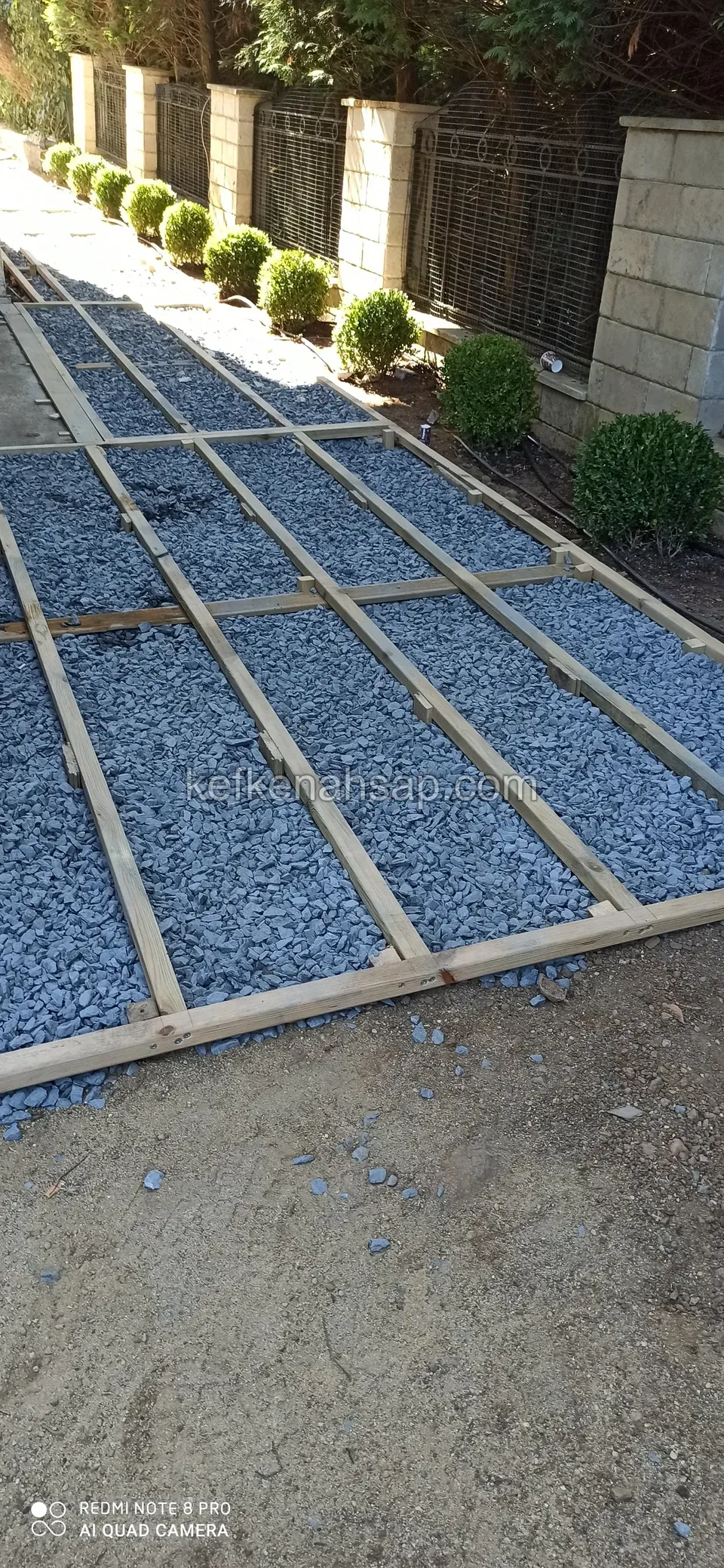 Pale timber deck substructure over blue-grey gravel running along a fence and wall