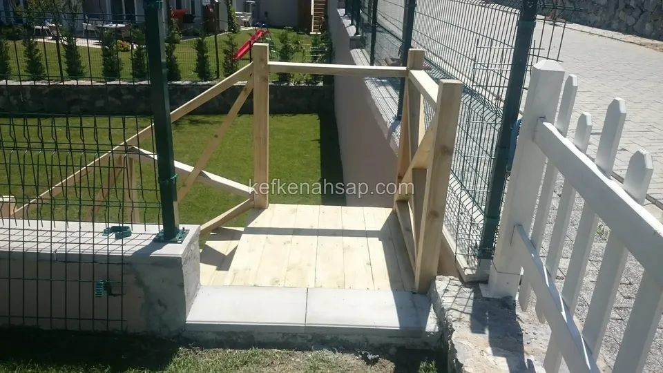 Beyaz çıtalı çit ve tel çit kenarında küçük ahşap platform; çaprazlı korkuluk, beton basamak ve çimlik