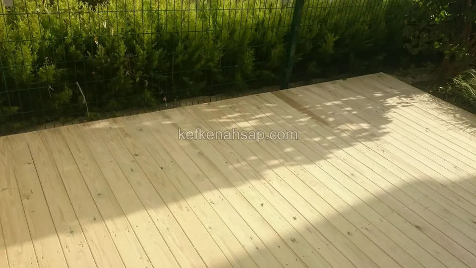 Telörgü çit ve çalı sırasının önünde, güneş ışığı alan açık renkli yeni deck yüzeyi