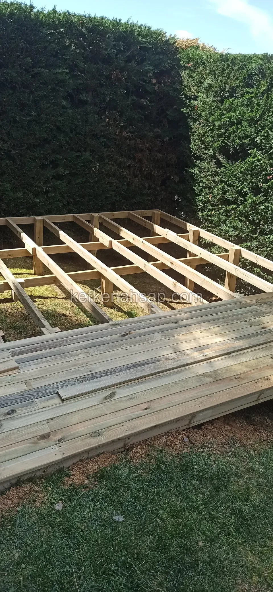 Çit önünde, ön kısmı tahtalarla kaplanmış, arka kısmında kirişleri açıkta duran deck, altta çimen