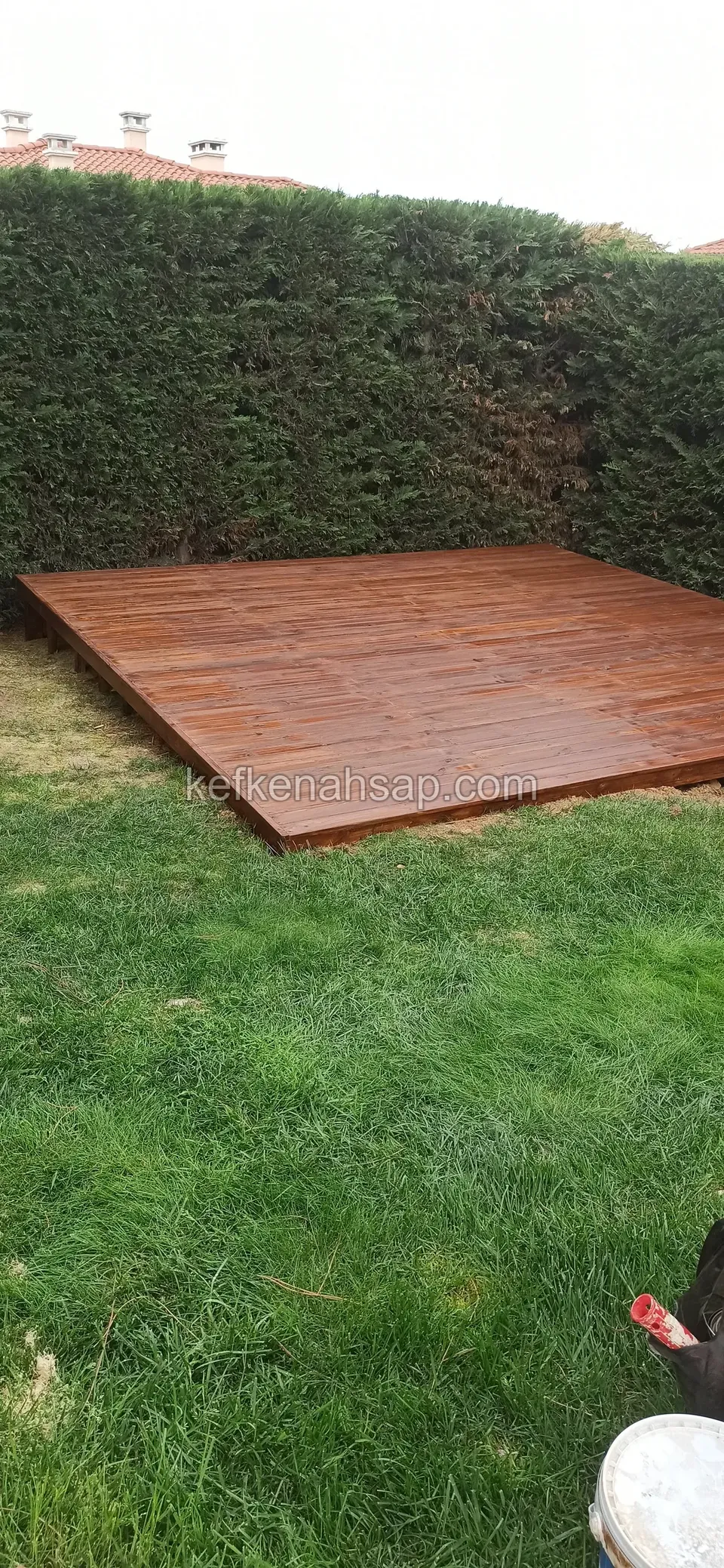 Brown stained deck laid on the lawn in front of a hedge, with an object partly visible in the lower right corner