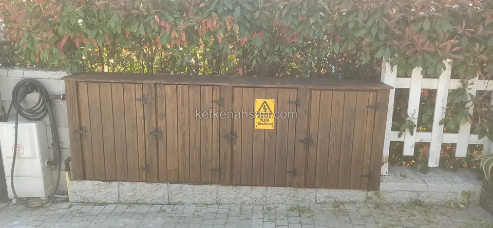 Çalı ve beyaz tahta çitin önünde taş tabanlı uzun koyu ahşap dolap, siyah menteşeli kapaklar ve sarı uyarı levhası