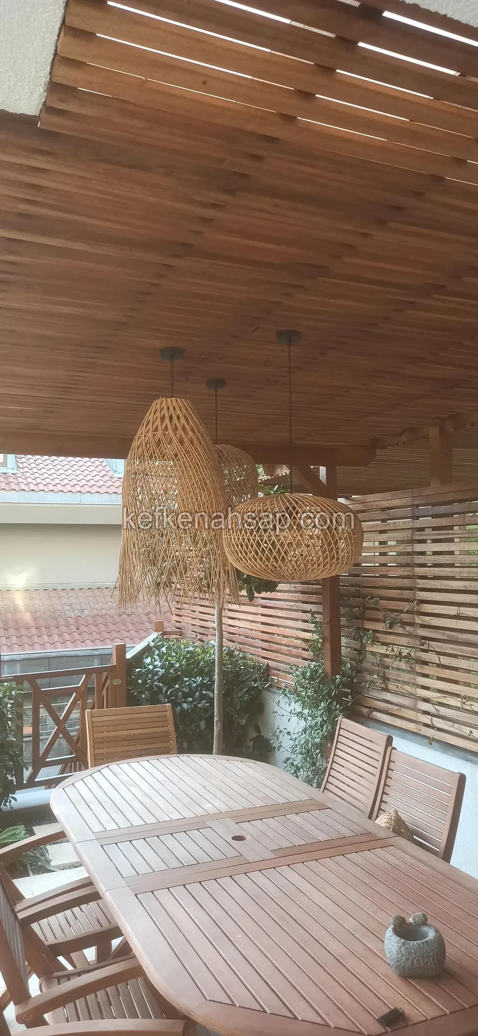 Çıtalı ahşap tavanın altında iki hasır avize, oval ahşap yemek masası ve sandalyeler, yanda çıtalı bölme