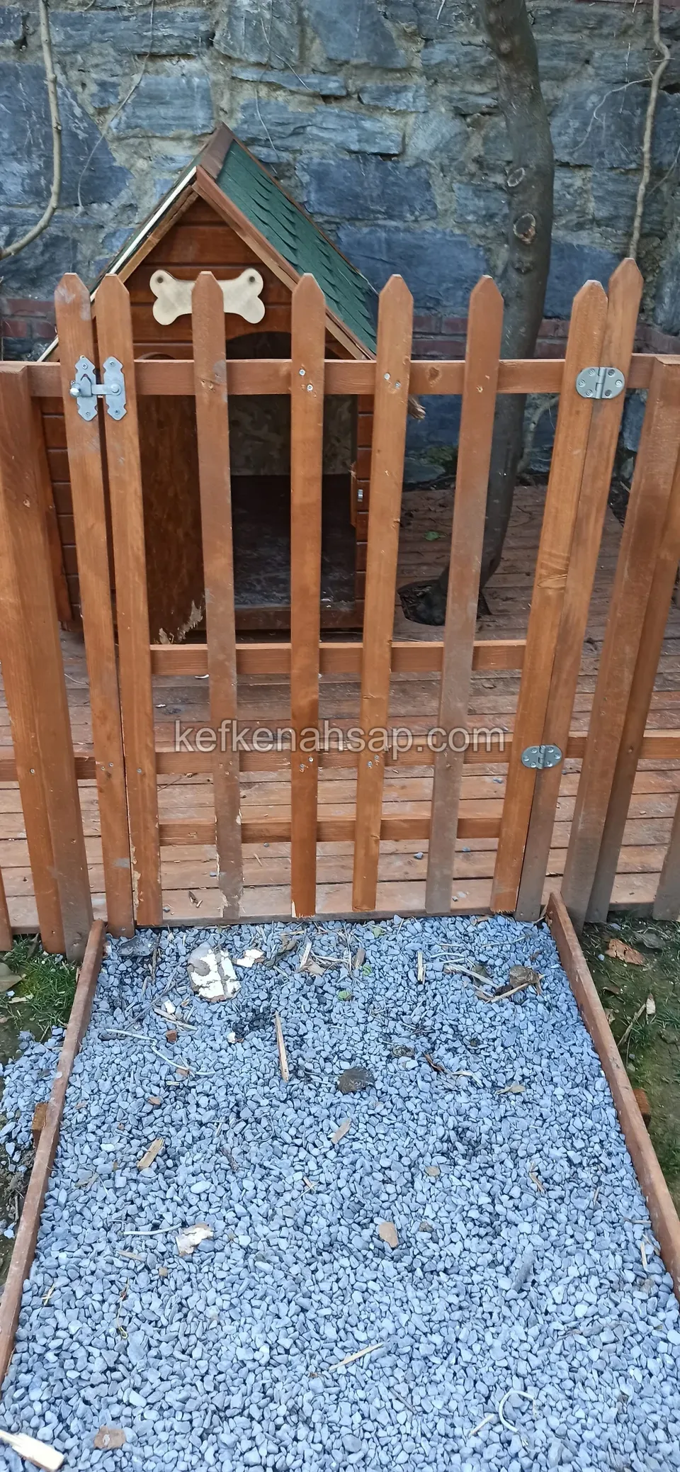 Sivri çıtalı ahşap kapının arkasında, çatısında kemik motifi olan küçük köpek kulübesi; önünde çakıl döşeli alan var