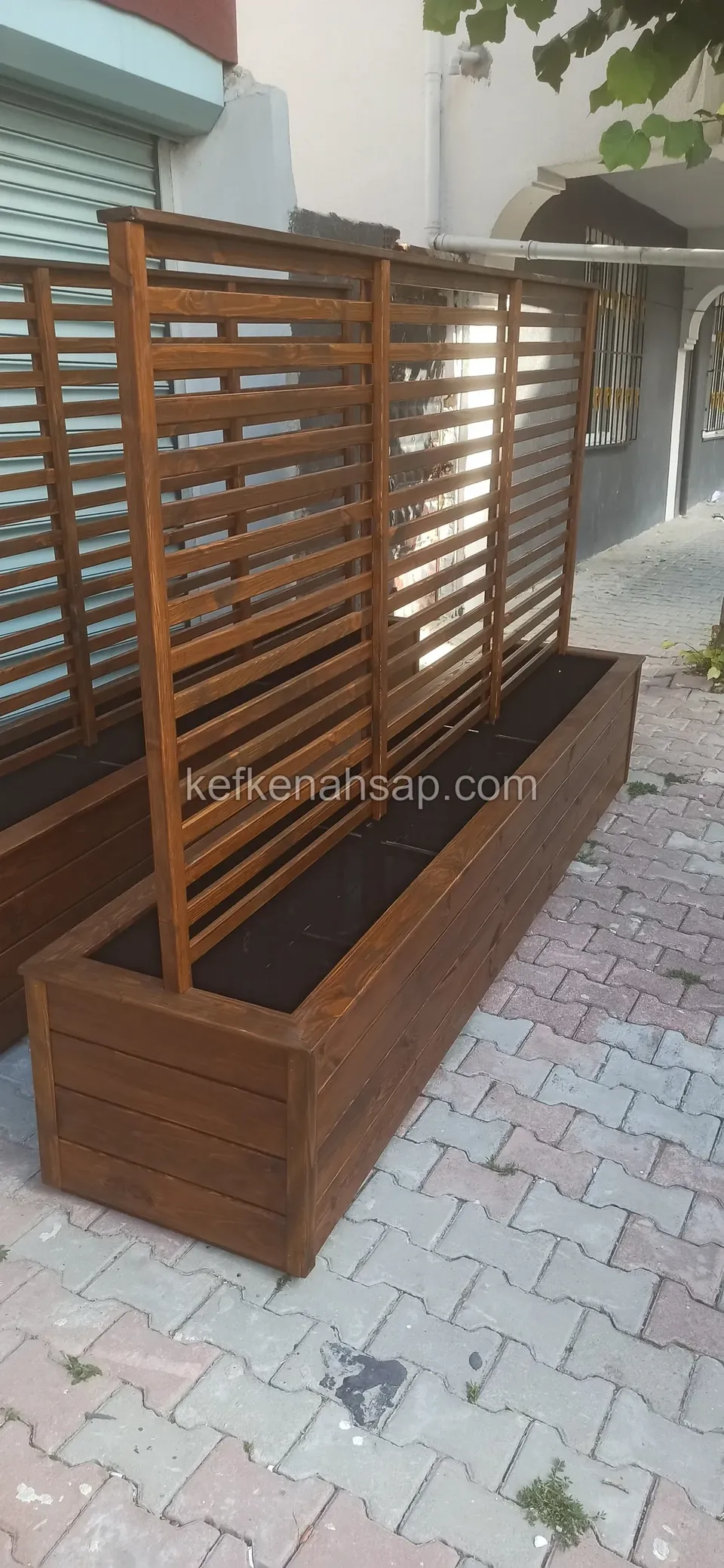 Kaldırımda duran, arkasında yatay çıtalı yüksek ahşap panel bulunan koyu cilalı dikdörtgen ahşap çiçeklik; toprağı henüz boş