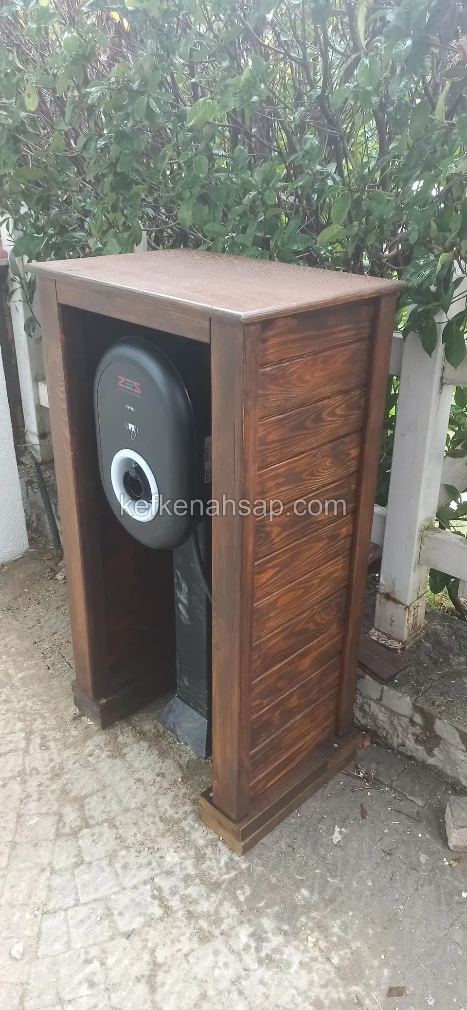 Parke taşlı zeminde yan yüzü yatay çıtalı, önü açık koyu ahşap kabin, içinde siyah yuvarlak bir cihaz