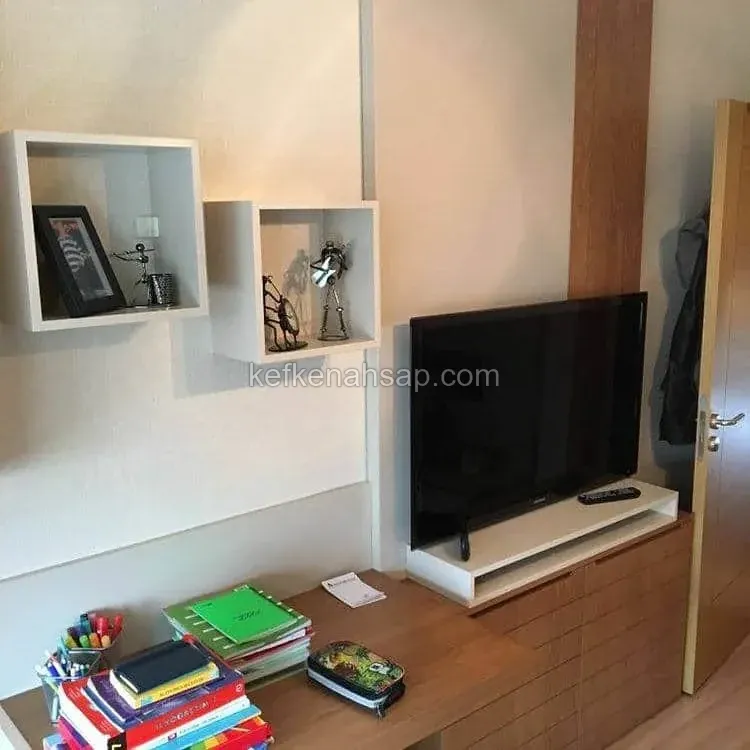 Beyaz kare duvar rafları, ahşap çekmeceli TV ünitesi, üzerinde televizyon ve çalışma tezgâhında kitaplar