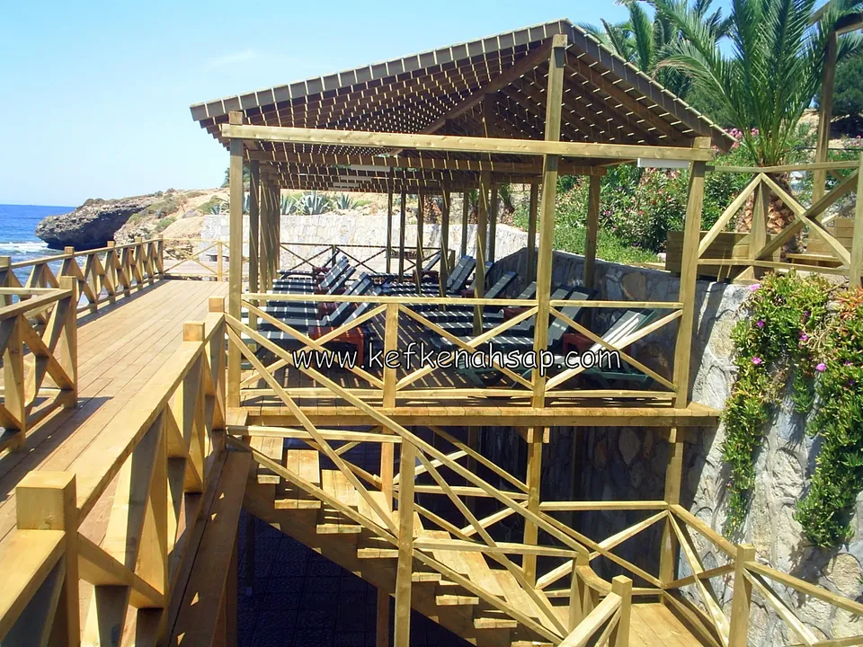 Mehrstufige Holzpergola mit Kreuzstreben-Geländer, Treppe und einer Reihe Sonnenliegen darunter, mit Meerblick