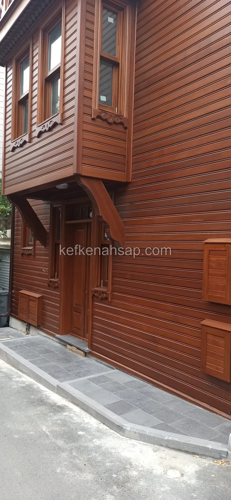 Cumbalı bir evin cephesinde kırmızımsı kahve tonlu vernikli yatay ahşap kaplama ve ahşap giriş kapısı, önde gri taş kaldırım