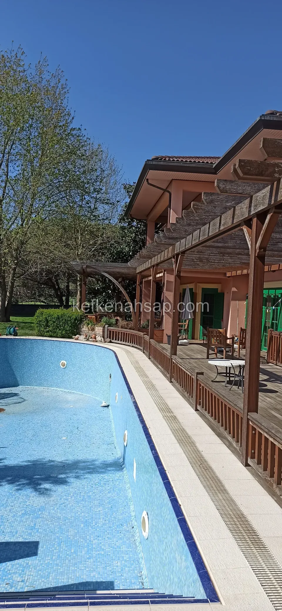 Dar ve uzun bir havuzun kenarında ahşap korkuluklu deck ve pergola, arkada kırmızı çatılı büyük ev