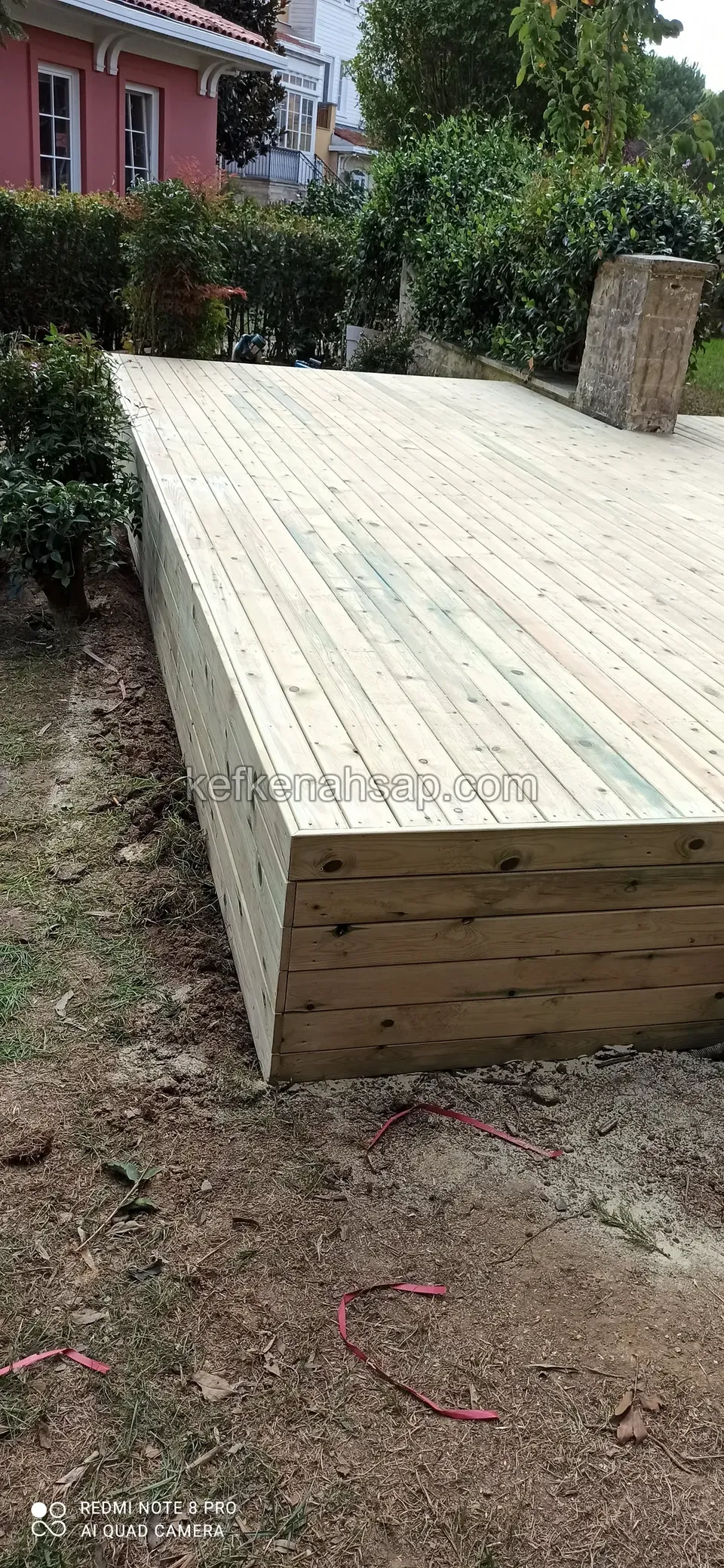Bahçede dar ve uzun yükseltilmiş deck platformu; yan yüzeyi yatay tahtalarla kaplanmış