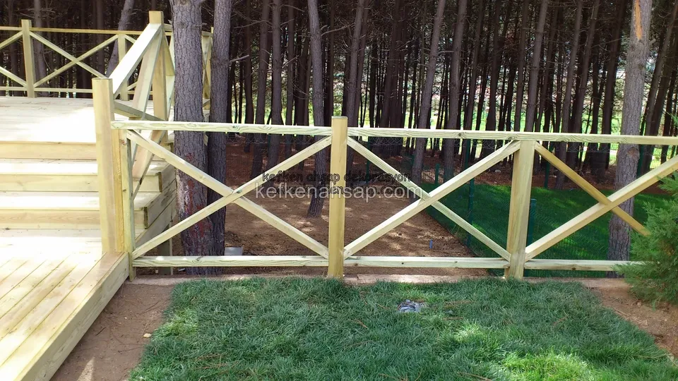 Çam ormanının içinde, açık tonlu deck platformun kenarında X çaprazlı ahşap korkuluk ve yeni serilmiş çim