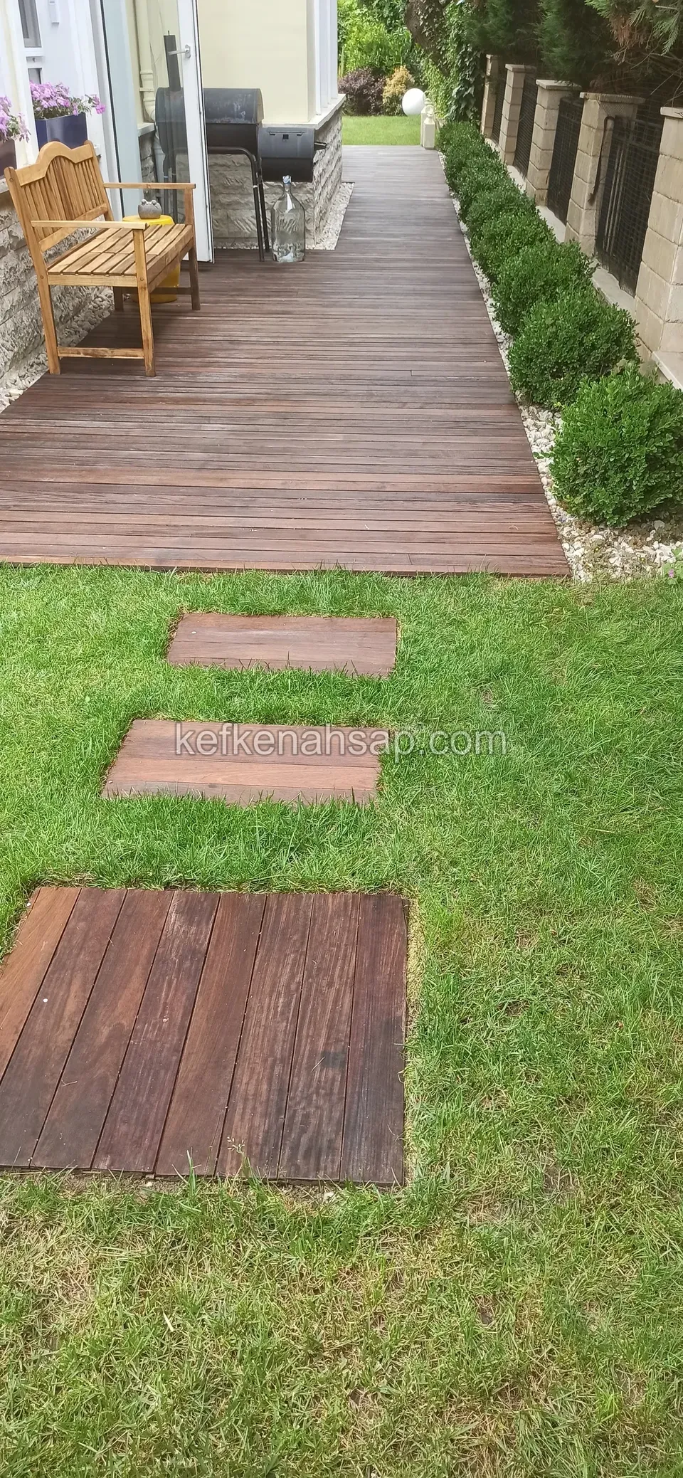 Çimde sıralı koyu basamak plakaları; uzakta deck üzerinde ahşap bank ve mangal, yanda kesme çalı sırası