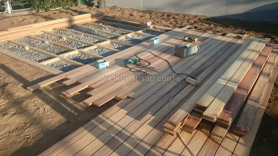 Half-laid timber deck over a gravel substructure, with a pile of boards and power tools beside it