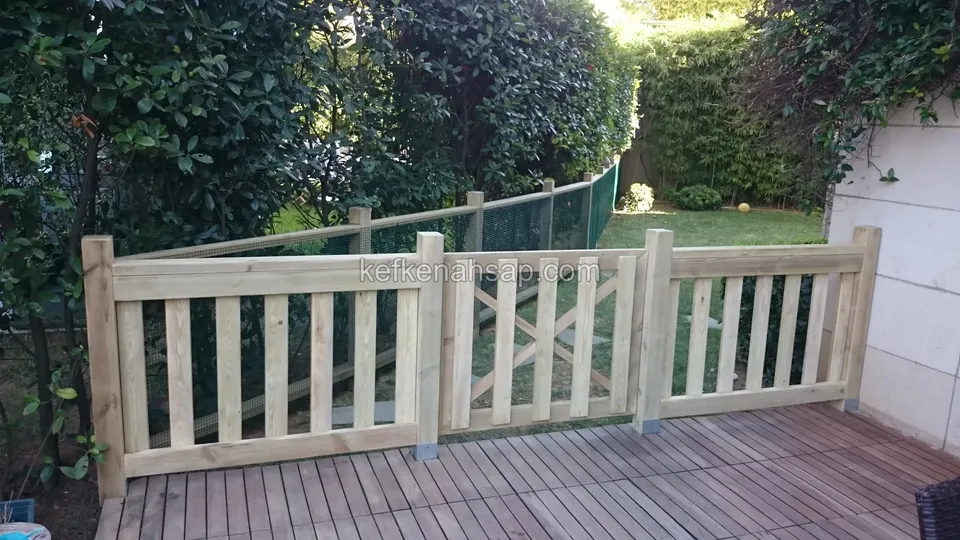 Karartılmış tahtalı deck zeminin kenarında ortası çaprazlı kapı olan üç bölmeli açık tonlu çıtalı korkuluk, arkada tel çit