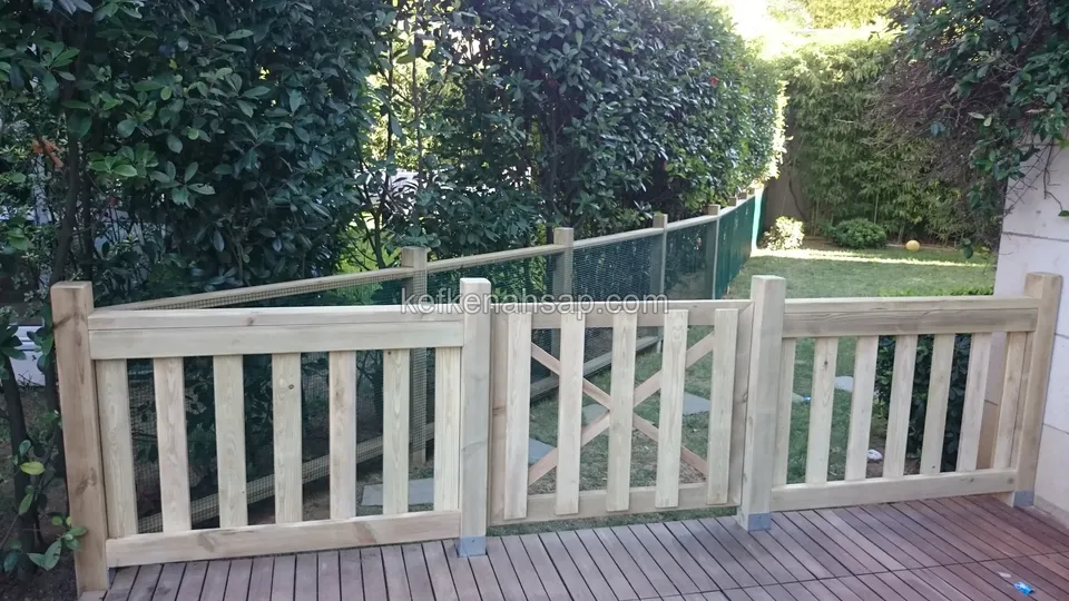 Koyu tonlu deck zeminin kenarında üç bölmeli açık tonlu çıtalı korkuluk, ortasında çaprazlı kapı ve arkasında yeşil tel çit