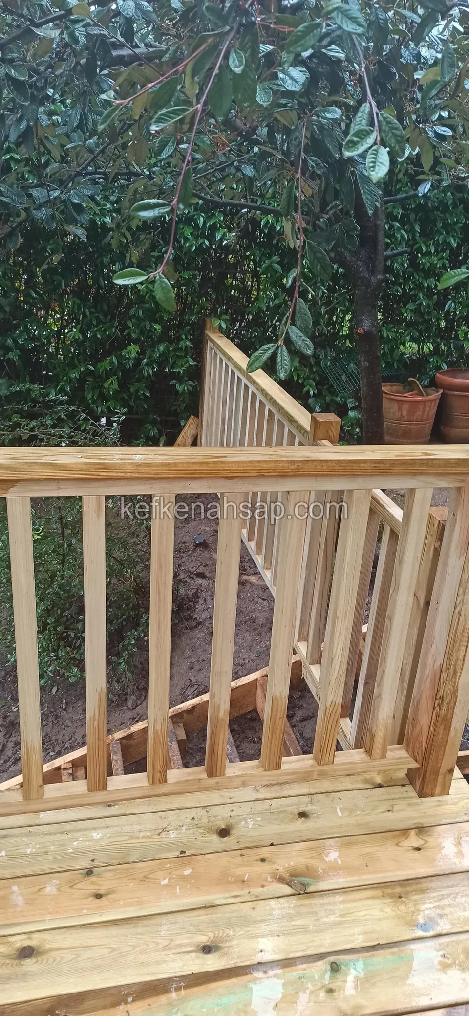 Açık tonlu yeni deck zemininin kenarında dikey çıtalı ahşap korkuluk, arkada ağaç yaprakları