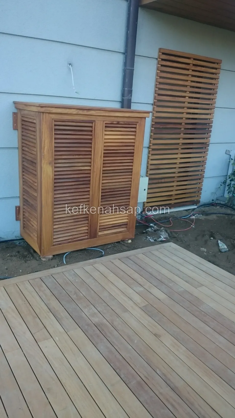 Mavi boyalı duvarın önünde deck zeminde duran jaluzi kapaklı küçük ahşap dolap ve yanında duvara yaslanmış çıtalı panel