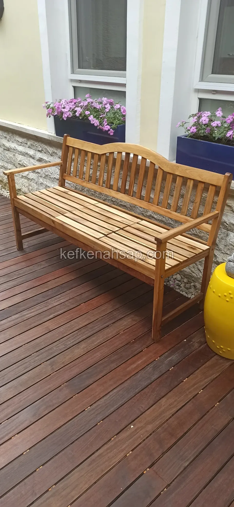 Çiçekli saksıların önünde, koyu tonlu ahşap deck üzerinde kollu çıtalı ahşap bank