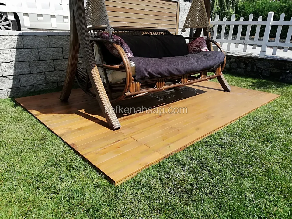 Çimen üzerine serilmiş ahşap deck ve üzerinde koyu minderli ahşap bahçe salıncağı
