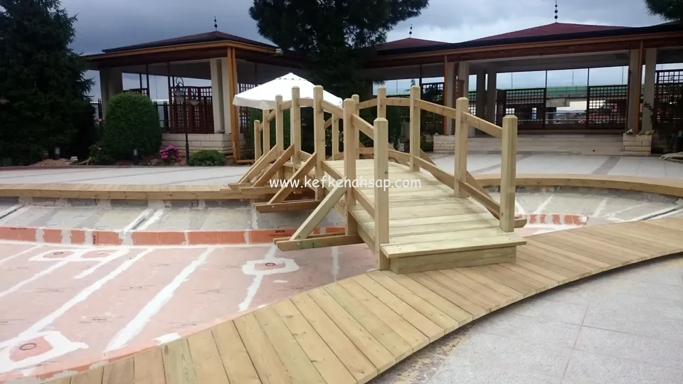 Kıvrımlı deck yolunun ucundan boş havuzu geçen korkuluklu ahşap köprü; arkada çatılı köşk ve şemsiye