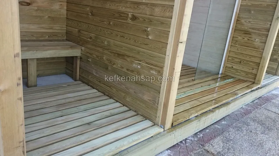 Yatay tahtalarla kaplı açık ahşap kabinin içinde duvara bağlı bir bank, zeminde aralıklı deck tahtaları var