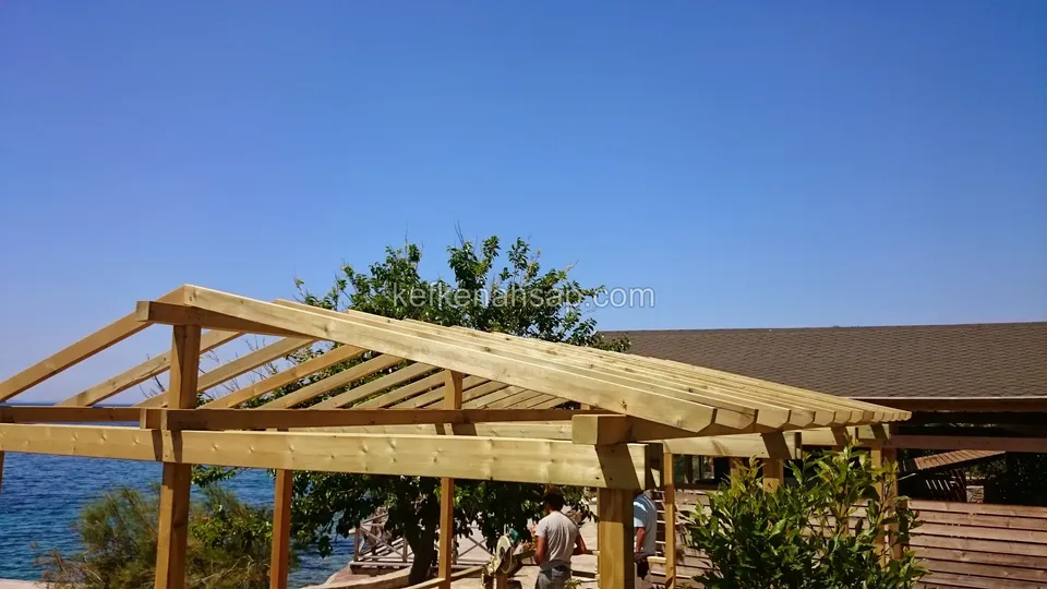 Deniz karşısında beşik çatılı ahşap pergola iskeleti, altında çalışan insanlar ve arkada kayalık kıyı