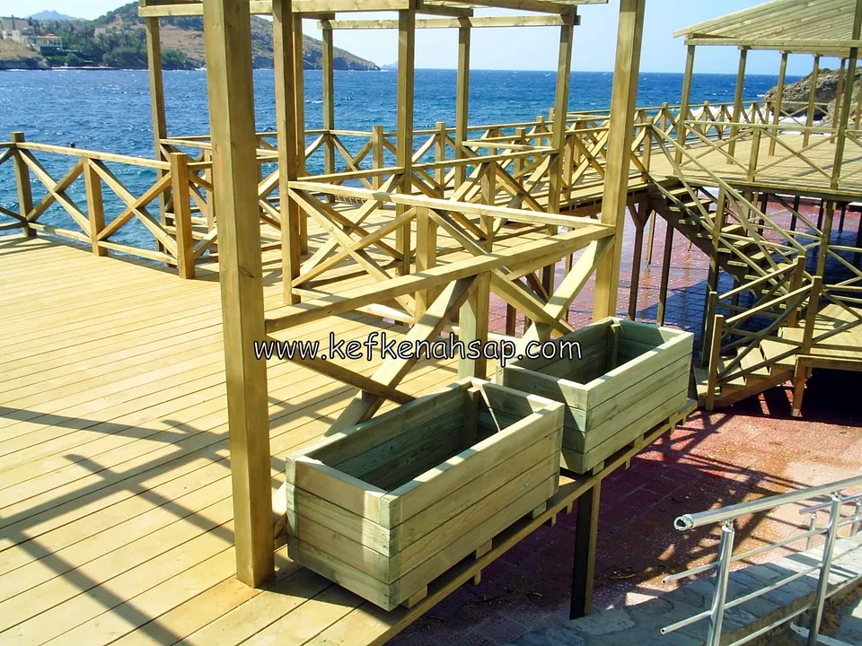 Denize bakan ahşap deck'in kenarında, çapraz korkulukların önünde duran iki dikdörtgen ahşap çiçeklik