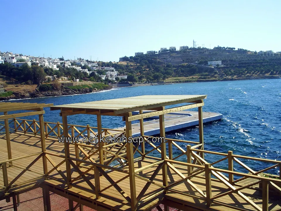 Dalgalı denizin kenarındaki deck üzerinde çapraz çıtalı korkuluk ve düz çatılı bir pergola, uzakta beyaz evler