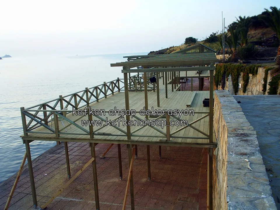 Taş duvara yaslanan, ince ayaklar üzerinde denize uzanan ahşap deck ve çevresinde X çaprazlı korkuluk, ucunda çatı kirişleri
