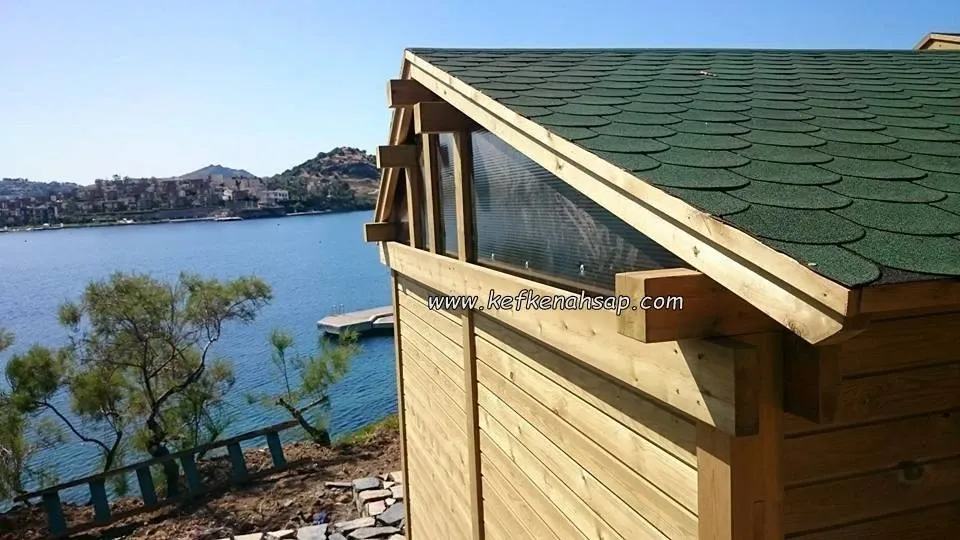Yeşil shingle çatılı, yatay kaplamalı ahşap yapının köşesi; arkada koy, yat iskelesi ve tepedeki yerleşim görünüyor