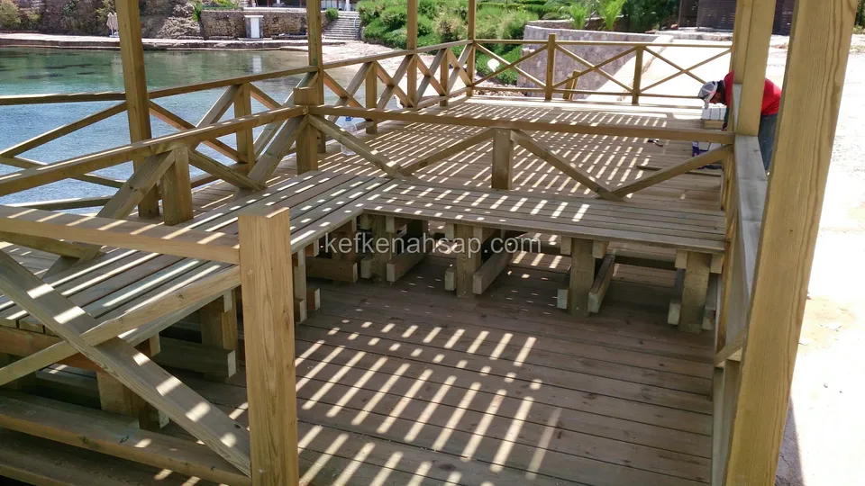 Deniz kıyısında çapraz çıtalı korkuluklu deck; içeride U şeklinde uzun ahşap bank ve masa