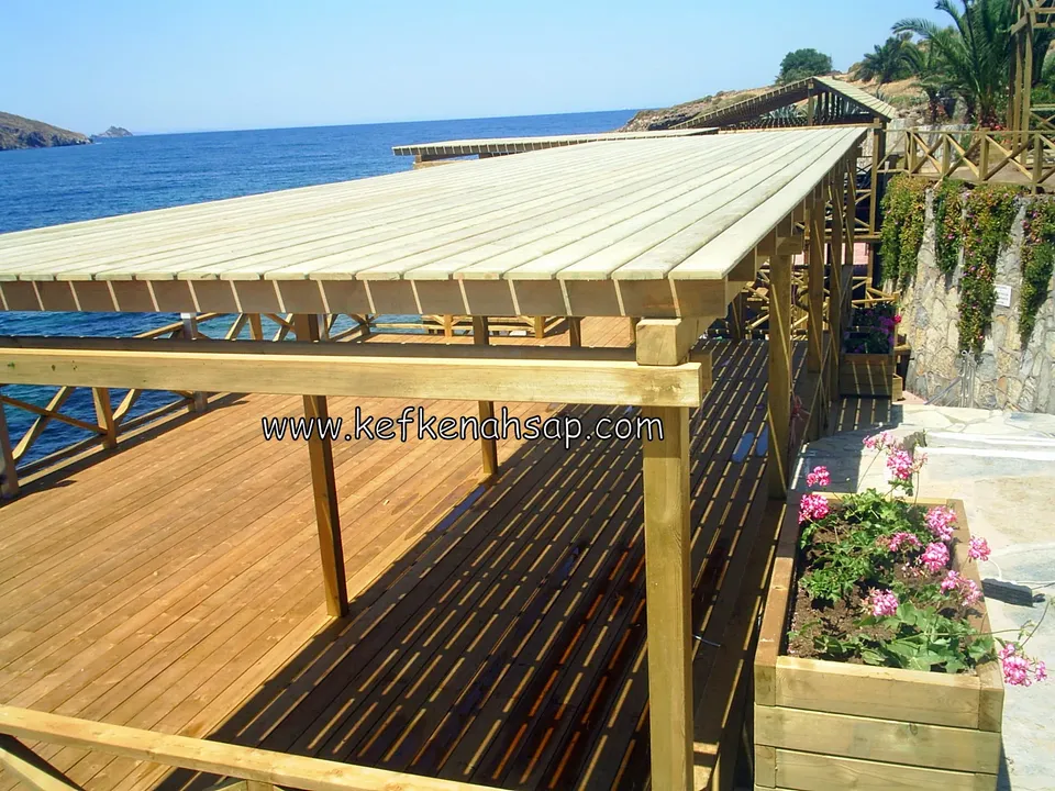 Deniz kenarındaki deck üzerinde tahta çatılı pergola ve ön tarafta çiçek açmış ahşap saksı