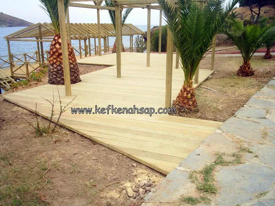 Deniz kenarında palmiyelerin arasında açık renkli ahşap deck; arkada ahşap pergola direkleri