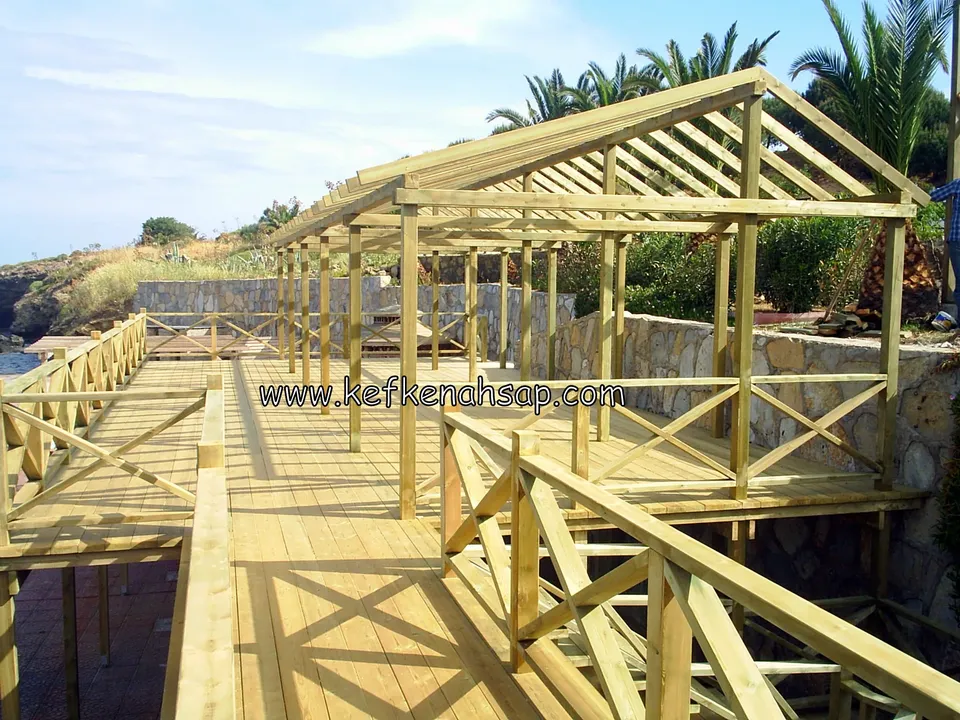 Deniz kenarında uzanan çatılı ahşap pergola, ahşap deck ve çapraz çıtalı korkuluk, palmiyeler