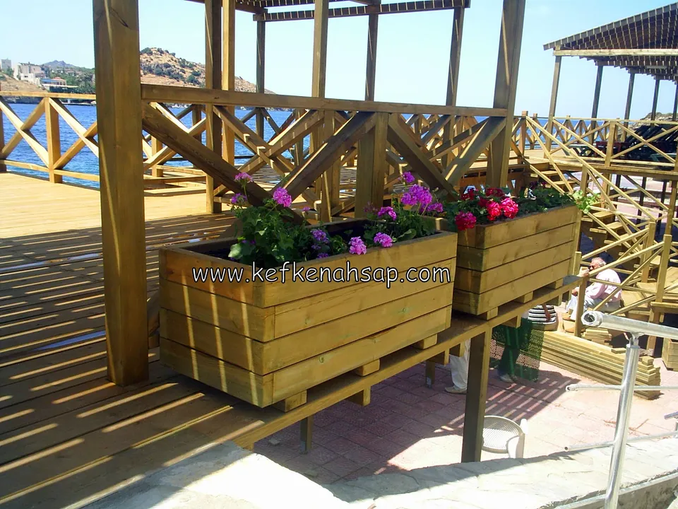 Two yellowish timber planters with pink and red geraniums on a timber deck by the sea