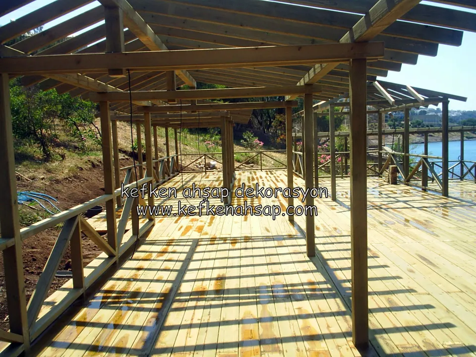 Denize bakan yamaçta, ahşap kolonlar ve kirişlerle yapılmış çatılı uzun pergola; zeminde deck, kenarlarda çapraz korkuluk var