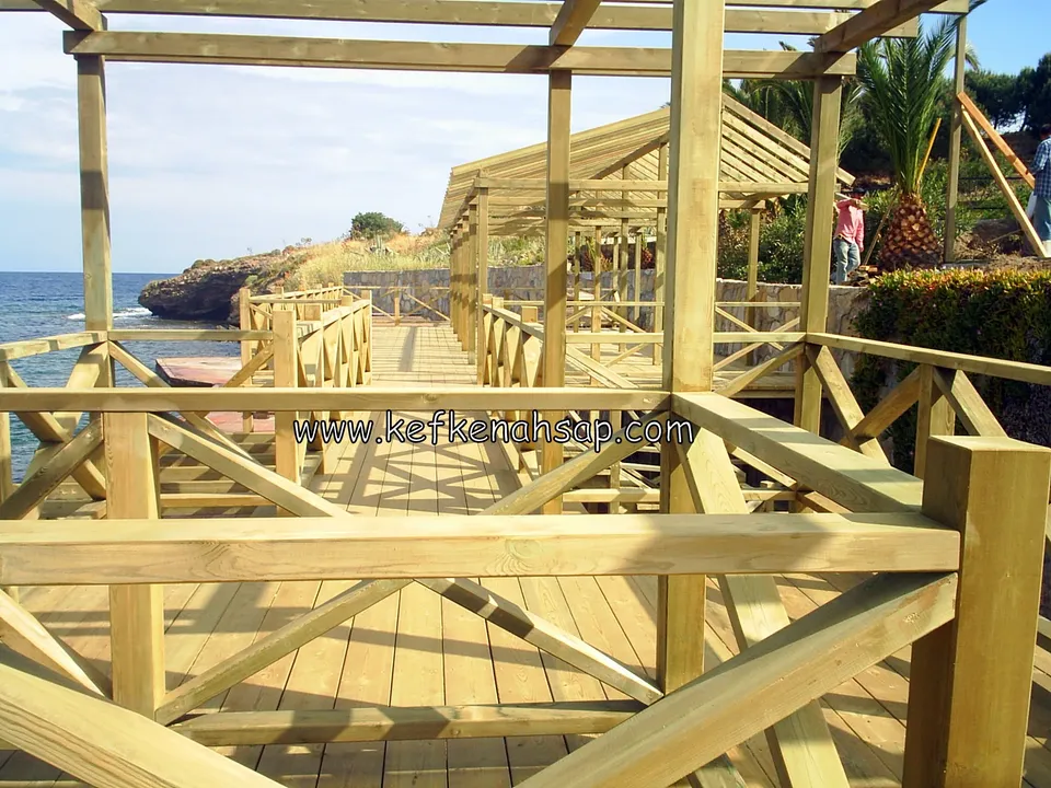 Denize bakan deck üzerinde kalın çaprazlı ahşap korkuluk, üstte pergola kirişleri, uzakta başka bir çatılı yapı ve palmiyeler