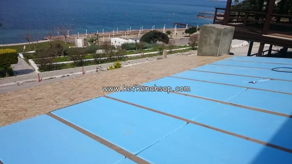 Deniz manzaralı bir çatı yüzeyinde ahşap çıtalar arasına döşenmiş mavi yalıtım levhaları, sağda ahşap bir yapı