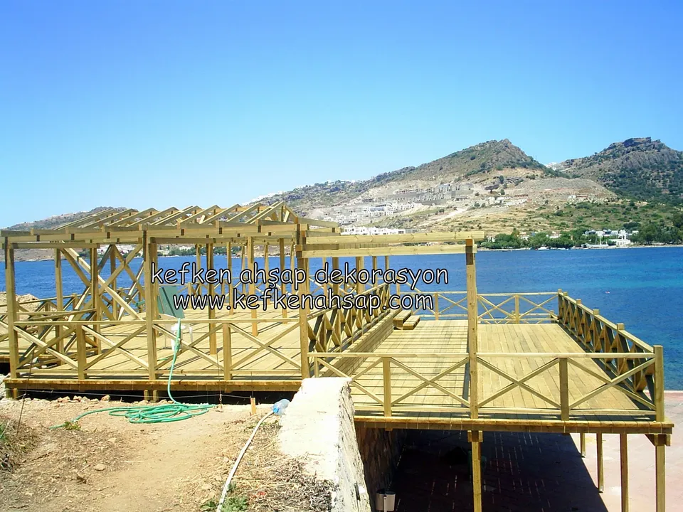Denizin hemen kıyısında, ayaklar üzerinde yükselen çapraz korkuluklu geniş deck; yerde hortum, arkada dağ silueti