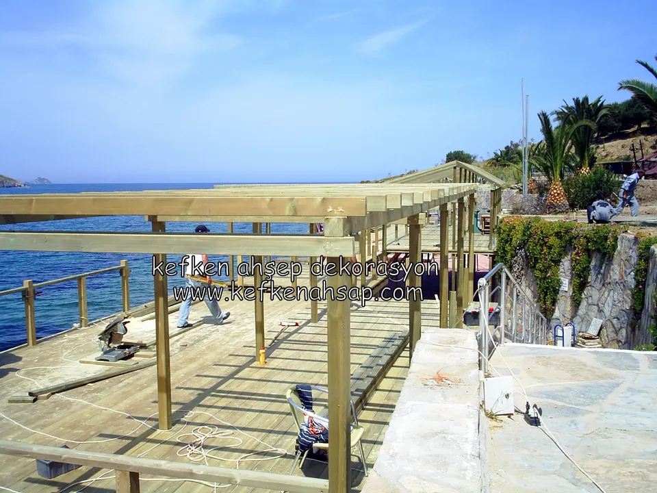 Deniz kıyısındaki deck üzerinde yükselen pergola iskeleti, çatı kirişleri takılıyor, işçiler çalışıyor