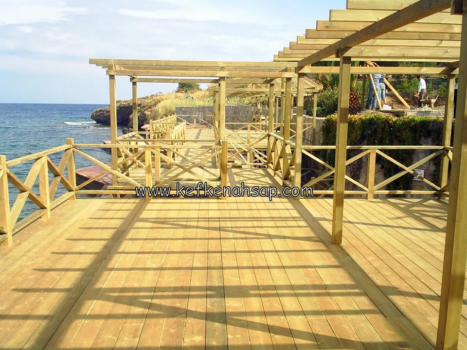 Deniz kıyısında çapraz X korkuluklarla çevrili, üzeri pergola çatılı geniş açık renkli deck, güneşte uzun gölgeler