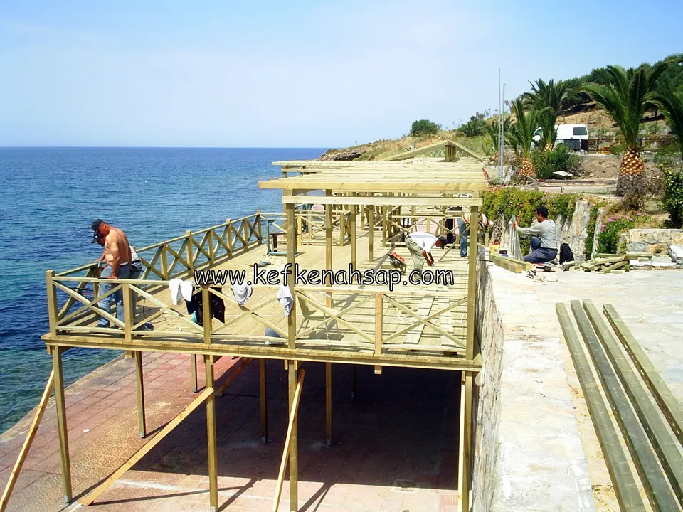 Denize bakan ahşap platformda çapraz çıtalı korkuluk ve pergola iskeleti; işçiler çalışıyor, sağda kalaslar duruyor