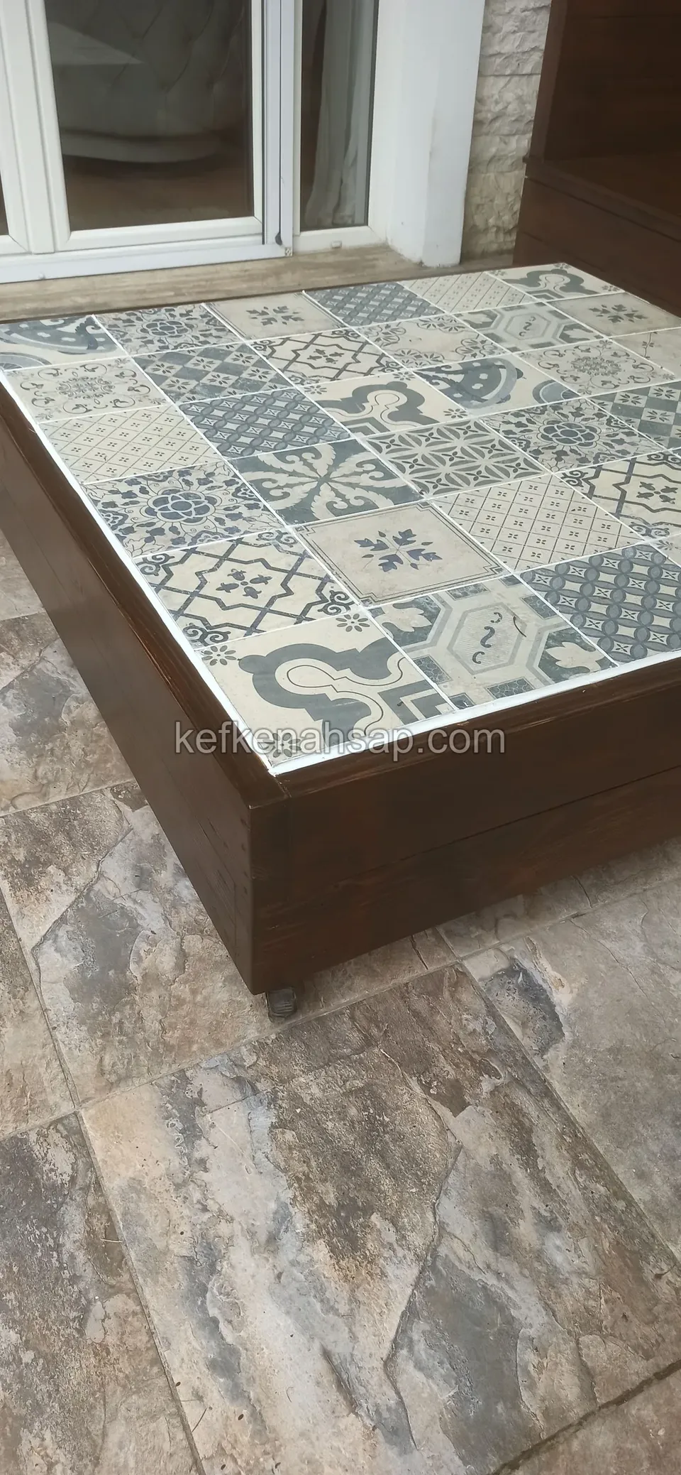 A low dark timber-framed table topped with blue-and-white patterned ceramic on stone paving beside a glass door
