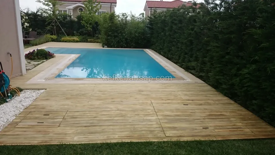 Dikdörtgen havuzun çevresini saran geniş açık renkli deck; yanında yüksek yeşil çit ve beyaz çakıl şeridi