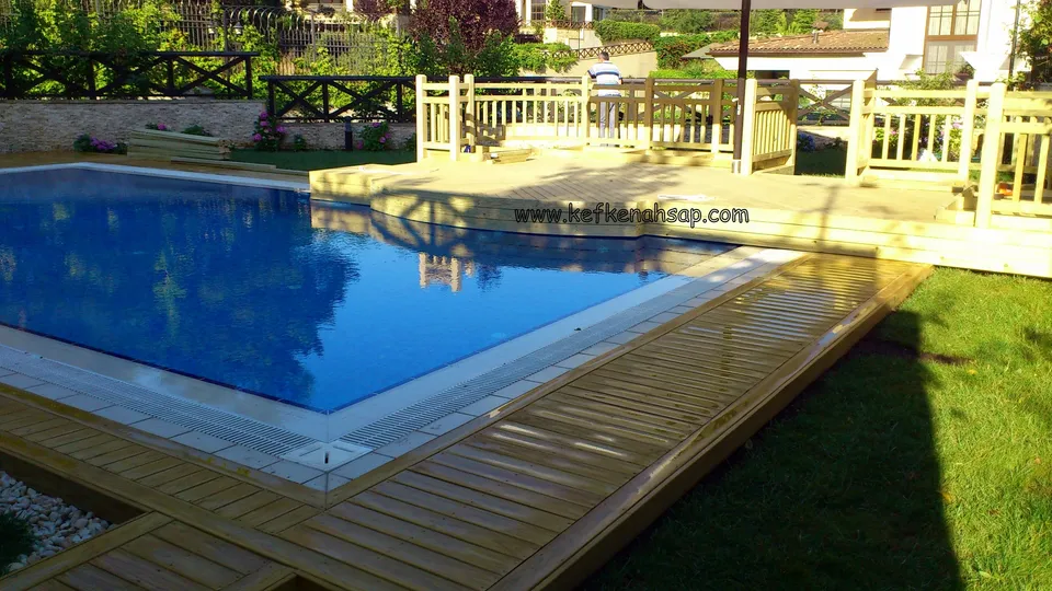 Mavi dikdörtgen havuzun kenarında uzanan açık renkli deck şeridi; arkada korkuluklu yükseltilmiş platform ve çimen