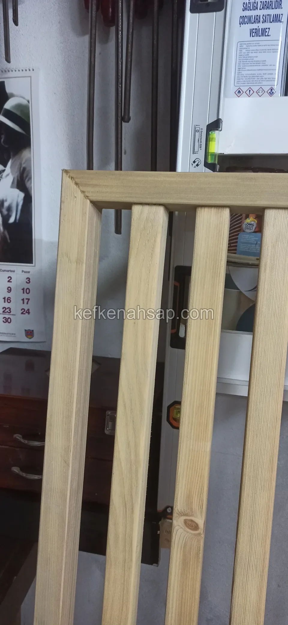 Nahaufnahme einer hellen Holztafel mit senkrechten Latten und oberem Rahmen vor einem unaufgeräumten Innenraum