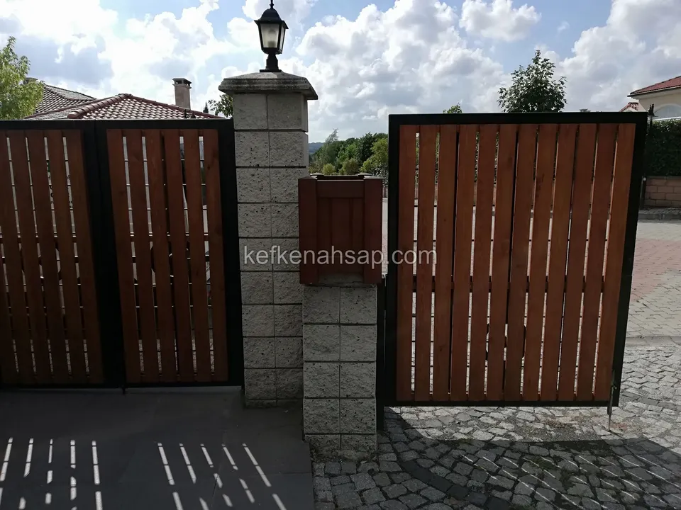 Küp taş döşeli yolda, siyah çerçeveli dikey çıtalı kahverengi kapı kanatları arasında lambalı taş sütun ve posta kutusu