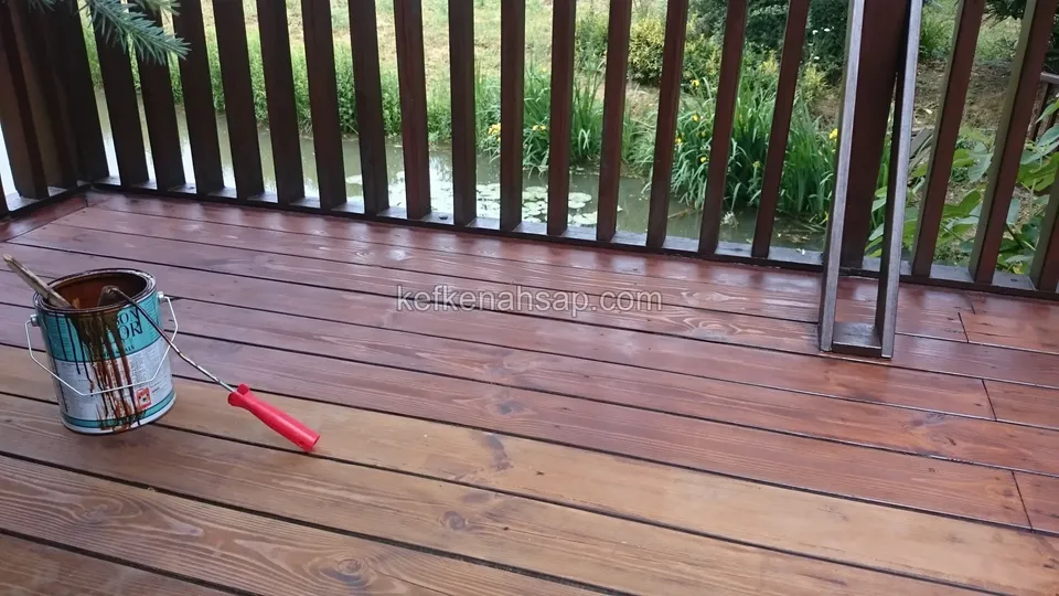 Deck being painted brown on a balcony with a vertical-slat timber railing, with an open paint tin